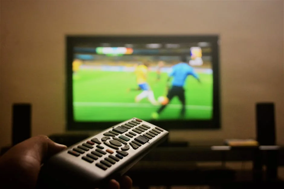 Close-up of a hand holding a remote control in front of a TV showing a football match. - m3u iptv
