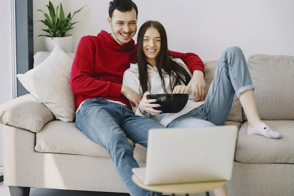 Cheerful man in casual wear embracing girlfriend with ceramic bowl while sitting on sofa and watching film together on netbook at home in sunlight - iptv subscriptions